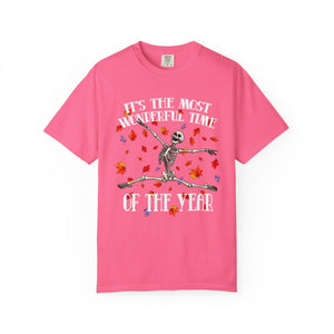 A pink short-sleeve crewneck t-shirt featuring a graphic of a skeleton dancing amidst colorful autumn leaves, with the text 'IT'S THE MOST WONDERFUL TIME OF THE YEAR' printed in white.