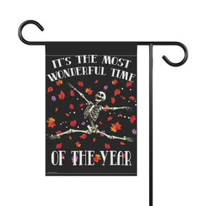 A black garden flag hanging from a black metal stand, featuring a skeleton sitting on a log with autumn leaves falling and the text 'IT'S THE MOST WONDERFUL TIME OF THE YEAR'.