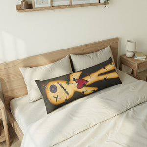 A long rectangular throw pillow with a stitched voodoo doll design rests on a bed with white linens and a light wood headboard in a softly lit bedroom.