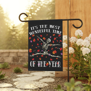 A decorative garden flag hanging from a black metal stand in a backyard setting. The flag features a skeleton dancing amidst colorful autumn leaves on a black background, with the text 'IT'S THE MOST WONDERFUL TIME OF THE YEAR'.