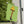 A green house flag mounted on a white exterior wall, featuring an illustration of a lamp shaped like a leg in fishnet stockings with a yellow lampshade and black high heel, set against a background of green trees and a beige house siding.