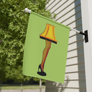 A green house flag mounted on a white exterior wall, featuring an illustration of a lamp shaped like a leg in fishnet stockings with a yellow lampshade and black high heel, set against a background of green trees and a beige house siding.