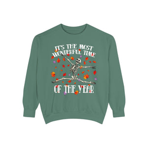 A sage green crewneck sweatshirt featuring a Halloween graphic of a dancing skeleton surrounded by falling autumn leaves, with the text 'IT'S THE MOST WONDERFUL TIME OF THE YEAR'.