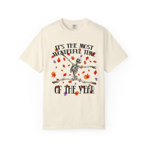 An off-white short-sleeve t-shirt with a graphic of a skeleton dancing amidst red and orange autumn leaves. The text reads 'IT'S THE MOST WONDERFUL TIME OF THE YEAR'.