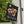 A black decorative flag hanging from a white pole on a house exterior, featuring cartoon characters under yellow and blue umbrellas above the word “FRIENDS” in the Friends from The Simpsons style. Trees and siding are visible in the background.