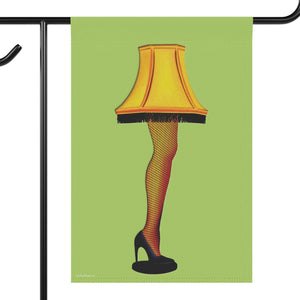 A garden flag hanging from a black metal stand, featuring a graphic illustration of a lamp shaped like a leg in fishnet stockings with a yellow lampshade and black high heels, set against a light green background.
