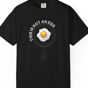 A black short-sleeve crewneck t-shirt featuring a graphic of a fried egg inside a frying pan with the text 'THIS IS NOT AN EGG' arched above it, displayed on a clean white background.