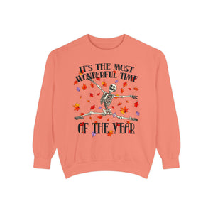 A coral pink crewneck sweatshirt featuring a Halloween graphic of a dancing skeleton surrounded by falling autumn leaves, with the text 'IT'S THE MOST WONDERFUL TIME OF THE YEAR'.