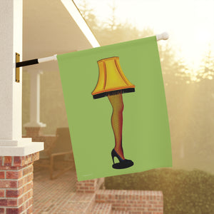 A decorative garden flag hanging from a white porch pole, featuring a stylized illustration of the Lamp of Guilty Thoughts: a yellow lampshade, fishnet stockings, and a black high heel on a light green background. The porch is softly blurred in the background with warm sunlight filtering through.