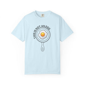A light blue short-sleeve crewneck t-shirt featuring a graphic of a fried egg inside a frying pan with the text 'THIS IS NOT AN EGG' arched above it, displayed on a clean white background.