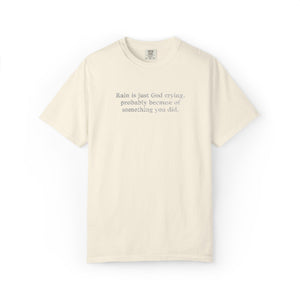 An off-white short-sleeve t-shirt with a printed quote on the chest that reads 'Rain is just God crying, probably because of something you did.' on a solid white background.