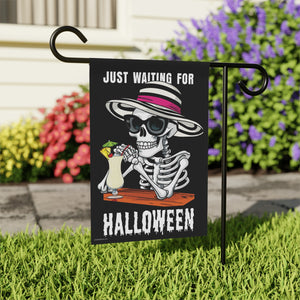 Yard flag with a skeleton wearing a hat and sunglasses drinking a pina colada and text that reads just waiting for summer, in a yard with flowers 