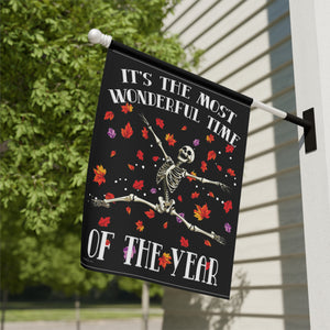 A black outdoor house flag hanging from a white wall mount, featuring a skeleton dancing among red, orange, and purple autumn leaves with the text 'IT'S THE MOST WONDERFUL TIME OF THE YEAR'.