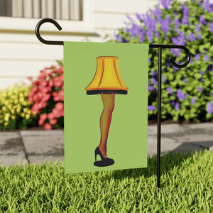 A rectangular garden flag hanging from a black metal stand in a grassy yard. The flag features an illustration of a yellow lampshade on a leg with fishnet stockings and a black high heel, set against a light green background.