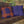 Brown swing with blue and orange pillows against a stone wall and greenery background