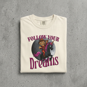 Folded t-shirt with a graphic of a person riding a unicorn and text 'Follow Your Dreams' on a concrete surface.