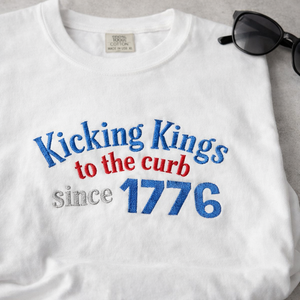 White t-shirt with 'Kicking Kings to the curb since 1776' text in blue and red on a gray background.