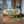A rectangular decorative pillow with a rustic wood-grain background, featuring the text 'Schweddy Balls' in green script with red berries and pine branches. The pillow is placed on a wooden table with a green ribbon tied to it, with a blurred kitchen background.