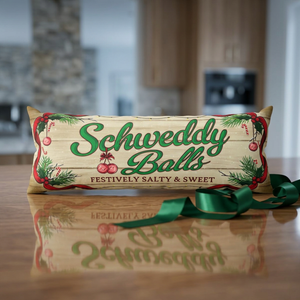 A rectangular decorative pillow with a rustic wood-grain background, featuring the text 'Schweddy Balls' in green script with red berries and pine branches. The pillow is placed on a wooden table with a green ribbon tied to it, with a blurred kitchen background.
