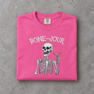 Folded pink t-shirt with a graphic of a skeleton holding a martini with Bone-Jour in bone style lettering at the top.