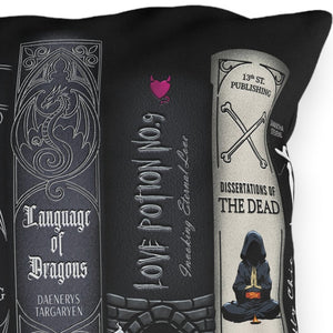 A square black throw pillow featuring a gothic-themed book spine print with illustrations and text such as 'Exorcisms,' 'The Art of Conjuring,' 'Language of Dragons,' 'Love Potion No. 9,' and 'Identification of the Dead.