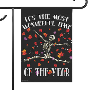 A black garden flag hanging from a black metal stand, featuring a dancing skeleton surrounded by colorful autumn leaves with the text 'IT'S THE MOST WONDERFUL TIME OF THE YEAR'.
