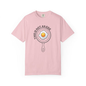 A light pink short-sleeve crewneck t-shirt featuring a graphic of a frying pan containing a fried egg with the text 'THIS IS NOT AN EGG' arched above it, displayed on a clean white background.