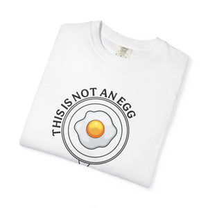 A folded white crewneck t-shirt featuring a graphic of a fried egg with the text 'THIS IS NOT AN EGG' in a circular layout, displayed on a clean white background.