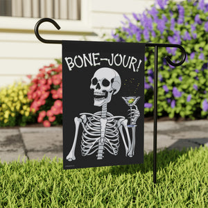 Yard flag and banner with a skeleton holding a martini that says Bone-jour at the top on a black background in a yard setting.