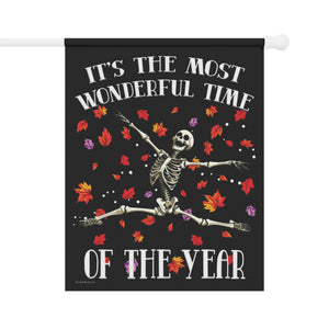 A vertical black decorative flag hanging from a white rod, featuring a dancing skeleton surrounded by red, orange, yellow, and purple autumn leaves. The text reads, 'IT'S THE MOST WONDERFUL TIME OF THE YEAR' in white lettering.