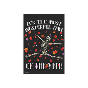 A rectangular black poster or sign featuring a white skeleton sitting cross-legged with the text 'IT'S THE MOST WONDERFUL TIME OF THE YEAR' in white arched lettering, surrounded by scattered red, orange, and purple autumn leaves.