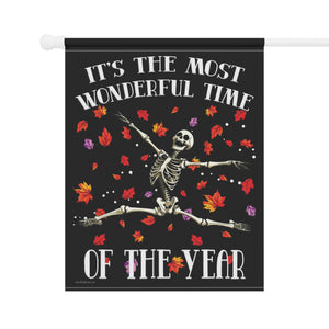 A vertical black decorative flag hanging from a white rod, printed with the phrase “IT’S THE MOST WONDERFUL TIME OF THE YEAR” in white arched letters around a dancing skeleton surrounded by red, orange, yellow, and purple autumn leaves.