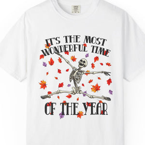 A white short-sleeve crewneck t-shirt featuring a Halloween graphic of a skeleton dancing amidst red, orange, and purple autumn leaves, with the text 'IT'S THE MOST WONDERFUL TIME OF THE YEAR'.
