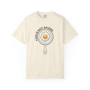 An off-white short-sleeve crewneck t-shirt featuring a graphic of a frying pan containing a fried egg with the text 'THIS IS NOT AN EGG' arched above it, displayed on a clean white background.