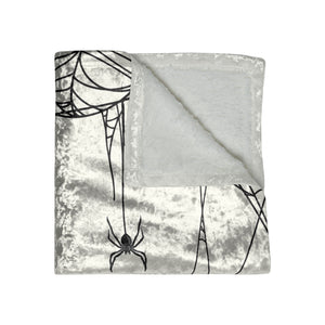 A square throw blanket with a silver-gray crushed velvet exterior featuring black spider and web illustrations, and a soft white lining, shown on a plain white background.