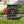 A black garden flag hanging from a black metal stand on a lawn, featuring a dancing skeleton surrounded by colorful autumn leaves with the text 'IT'S THE MOST WONDERFUL TIME OF THE YEAR'. The background shows a blurred outdoor garden with purple and yellow flowers.