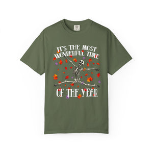 An olive green short-sleeve t-shirt with a graphic of a dancing skeleton surrounded by red and purple autumn leaves, with the text 'IT'S THE MOST WONDERFUL TIME OF THE YEAR' in white.