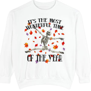 A white crewneck sweatshirt featuring a Halloween graphic of a skeleton dancing with a broom, surrounded by falling red and orange autumn leaves. The text reads 'IT'S THE MOST WONDERFUL TIME OF THE YEAR'.