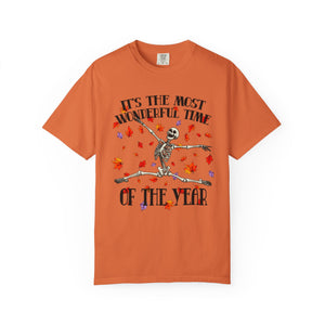 An orange short-sleeve crewneck t-shirt featuring a Halloween graphic of a dancing skeleton surrounded by falling red and purple leaves, with the text 'IT'S THE MOST WONDERFUL TIME OF THE YEAR'.
