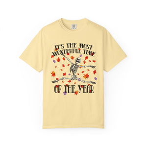 A pale yellow short-sleeve t-shirt with a graphic of a dancing skeleton surrounded by red and orange autumn leaves, with the text 'IT'S THE MOST WONDERFUL TIME OF THE YEAR'.