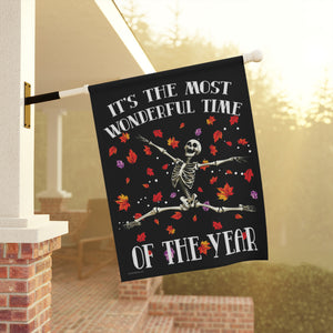 A black decorative garden flag hanging from a porch wall. The flag features a white skeleton dancing with colorful red, orange, and purple autumn leaves, with the text 'IT'S THE MOST WONDERFUL TIME OF THE YEAR' in white lettering.
