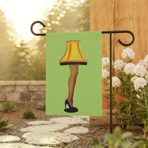 A rectangular garden flag hanging from a black metal stand in a garden. The flag features an illustration of the iconic 'This is fine' lamp from The Addams Family, with a yellow lampshade, red-and-black striped leg, and black high heel on a light green background.