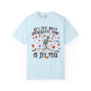 A light blue short-sleeve t-shirt with a Halloween-themed graphic on the front. The graphic features a skeleton dancing amidst red, orange, and purple autumn leaves, with the text 'IT'S THE MOST WONDERFUL TIME OF THE YEAR' in a stylized font.