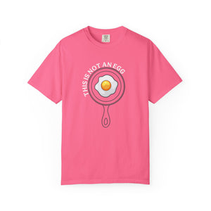 A pink short-sleeve crewneck t-shirt featuring a graphic of a frying pan with a fried egg inside and the text 'THIS IS NOT AN EGG' arched above it, displayed on a clean white background.