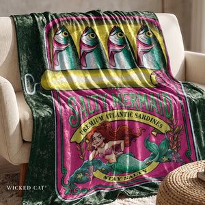 A plush throw blanket draped over a cream armchair, featuring a colorful 'Salty Mermaid' sardine can design with mermaid artwork and fish illustrations. The blanket has a dark green velvet-like border and a bright pink center panel.