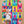 A baby in a white bodysuit stands on a colorful printed blanket featuring a repeating pop-art bottle design on a wood floor. The blanket has multiple bright colors and white paint-splatter motifs.