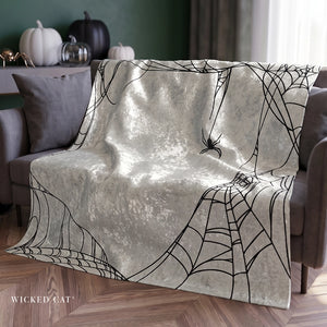 A plush throw blanket draped over a gray armchair in a styled living room. The blanket has a distressed, textured off-white background with a black spiderweb and spider print.