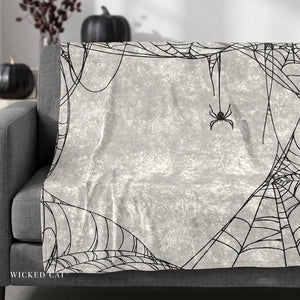A gray throw blanket draped over a gray sofa, featuring a black spider and intricate spiderweb pattern on a textured off-white background. The scene is styled with dark pumpkins in the background.