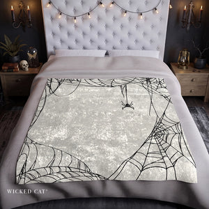 A bed dressed in gray bedding with a large printed blanket or throw featuring black spiderwebs and a spider on a textured off-white background. The bed is styled in a dark, moody bedroom with a tufted headboard, bedside tables, candles, and string lights.