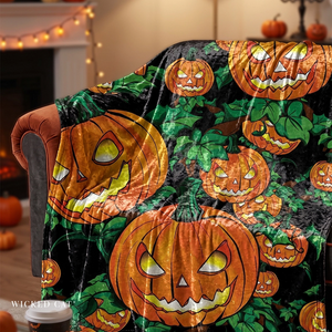 A Halloween-themed throw blanket draped over an armchair in a cozy indoor setting. The blanket features a repeating pattern of orange jack-o'-lantern pumpkins with glowing yellow eyes and mouths on a black background with green leaves.